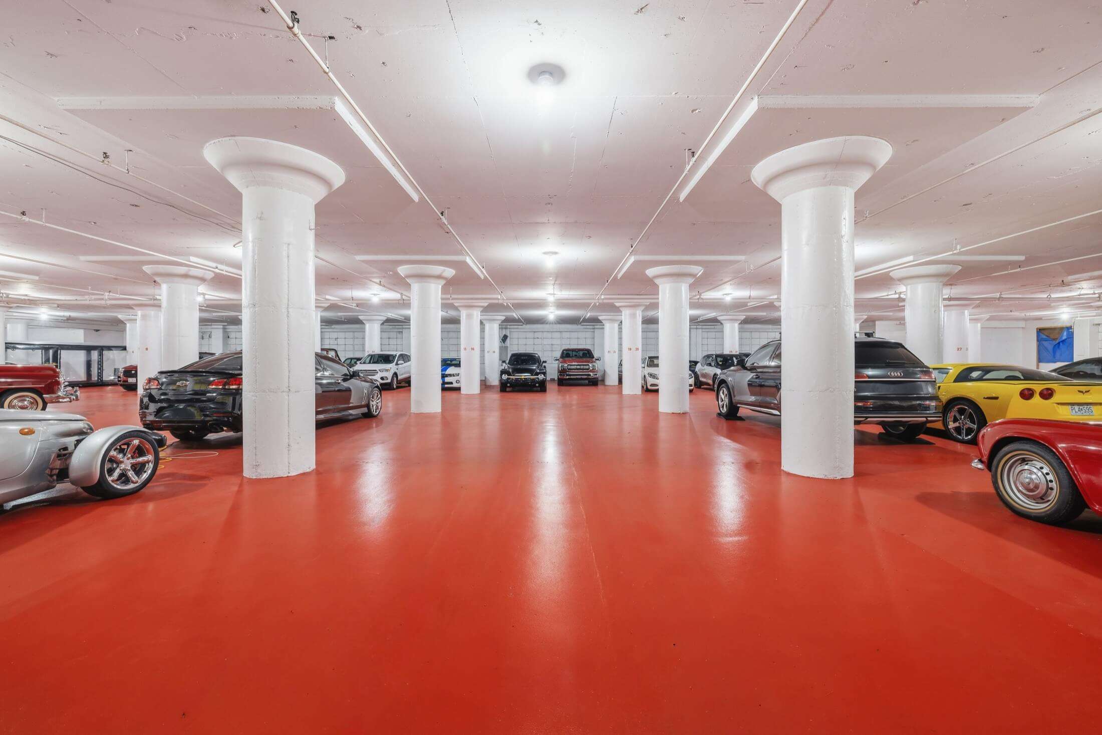 Indoor storage facility with vintage and modern cars parked alongside white support columns and bright red floor